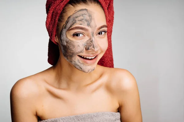 Giovane ragazza carina con un asciugamano rosso sulla testa sorridente, maschera di argilla mezza faccia, day spa — Foto Stock
