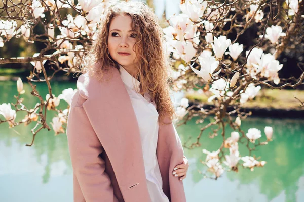 Curly-haired woman wearing a pink coat posing next to a blooming fragrant magnolia in a park, ranks with a pond — Stock Photo, Image