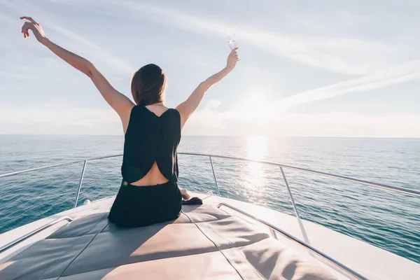 Молодая богатая девушка в черной летней одежде сидит на белой яхте на солнце, наслаждается морским путешествием, поднимает руки — стоковое фото