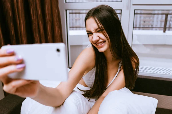 Divertida hermosa joven sentada en la cama temprano en la mañana y haciendo selfie, sonriendo —  Fotos de Stock