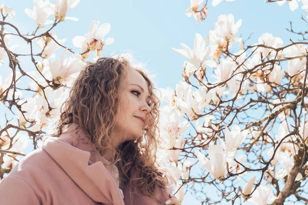 Mujer joven y rizada camina por el parque de primavera, disfruta de una magnolia floreciente fragante y el sol — Foto de Stock