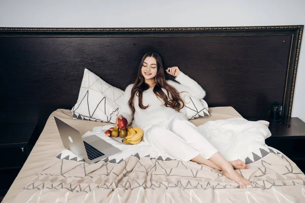 Ein nettes junges Mädchen im weißen Schlafanzug liegt auf einem Bett, isst nützliches Obst und schaut sich auf ihrem Laptop eine interessante Fernsehsendung an — Stockfoto