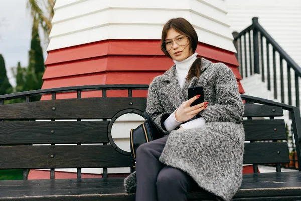Une fille urbaine aux cheveux bruns avec des lunettes et un manteau élégant gris s'assoit sur un banc et tient un smartphone dans sa main, son style de rue et sa mode — Photo