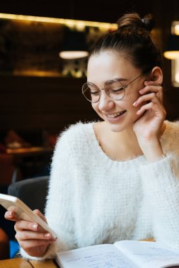 mutlu genç kız öğrenci gözlük ve beyaz bir ceket Üniversitesi'nde okuyan, bir akıllı telefon bakıyor ve gülümseyen sonra bir kafede oturuyor