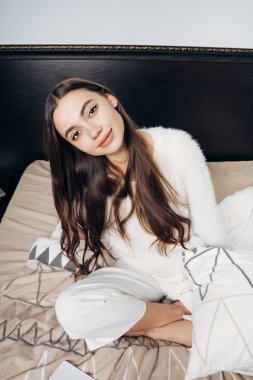 kameraya bakıyor ve gülümsüyor, güzel şirin kız yatakta oturup beyaz pijamalı yattım