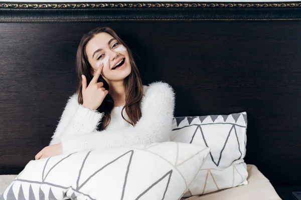 Una dulce chica somnolienta en pijama blanco yace en la cama, temprano en la mañana, bajo los ojos de manchas blancas, sonriendo — Foto de Stock