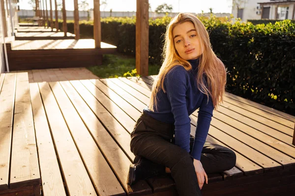 सुनहरे बालों वाली सुंदर प्यारी लड़की सूरज में एक पार्क में एक लकड़ी की बेंच पर बैठी — स्टॉक फ़ोटो, इमेज