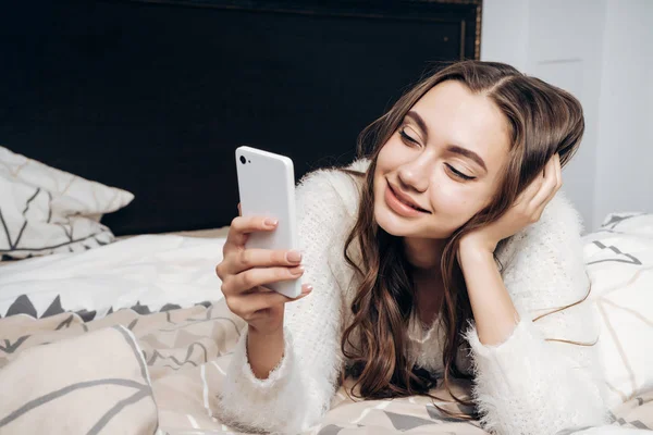Schattig jong meisje ligt op bed, kijkt naar haar smartphone en glimlacht — Stockfoto