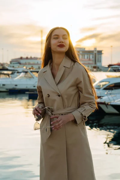 Красивая привлекательная девушка с длинными волосами и в модном бежевом пальто стоит в морском порту и улыбается — стоковое фото