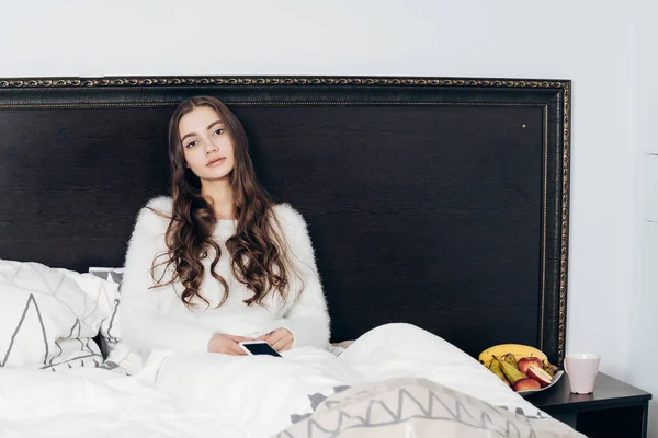 Beautiful sleepy girl in white pajamas sits in bed, resting after working day — Stock Photo, Image