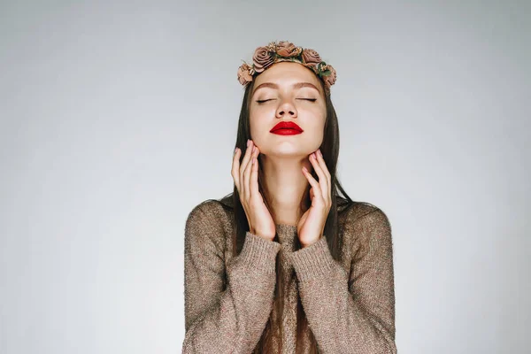 Una niña con lápiz labial brillante y en una hermosa corona de flores en la cabeza, cerró los ojos y levantó la cabeza, tocando suavemente su cara con las manos. estudio brillante — Foto de Stock