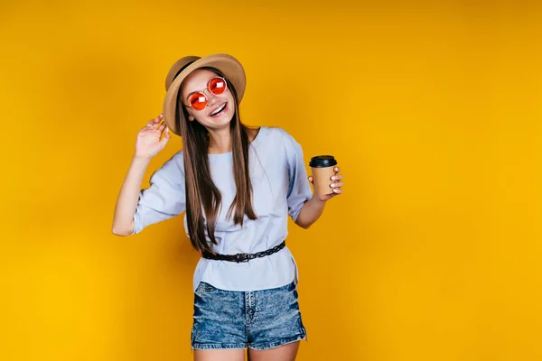 Estúdio amarelo. uma menina em um chapéu e óculos rosa detém café e alegremente caretas — Fotografia de Stock