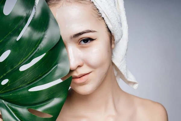 Een Aziatische vrouw in een handdoek op haar hoofd bedekt de helft van haar gezicht met een blad van monstera, haar schouders zijn kaal — Stockfoto