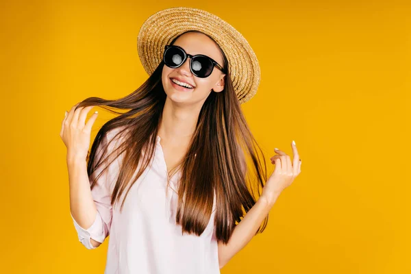 Chica con sombrero y gafas negras sonríe alegremente y toca el pelo con las manos sobre un fondo amarillo — Foto de Stock