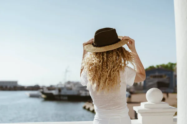 Dziewczyna z kręconymi blond włosami zakłada kapelusz na tle statków i morza. Dziewczyna stoi z tyłu — Zdjęcie stockowe
