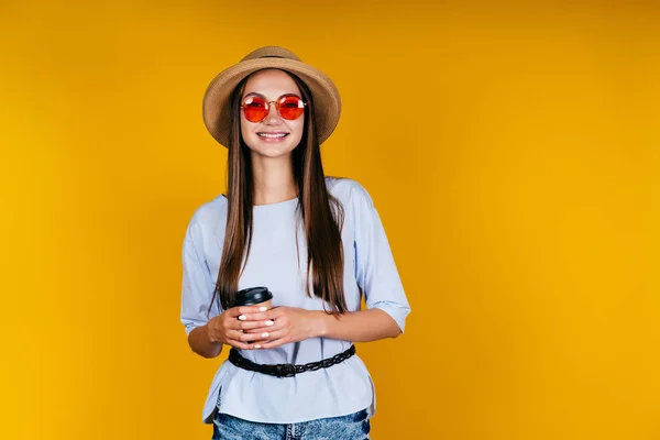 Studentessa in occhiali rosa e un cappello sorridente è in piedi in studio con un bicchiere di caffè. sfondo giallo — Foto Stock