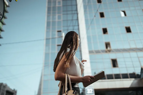 A student with a tablet pc in a half-turn hand stands against the backdrop of a huge glass building — Stock Photo, Image