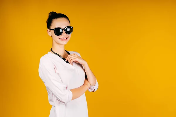 Una chica con un bulto en la cabeza y con gafas negras, sonríe y cubre con la mano el área debajo del pecho. fondo amarillo — Foto de Stock