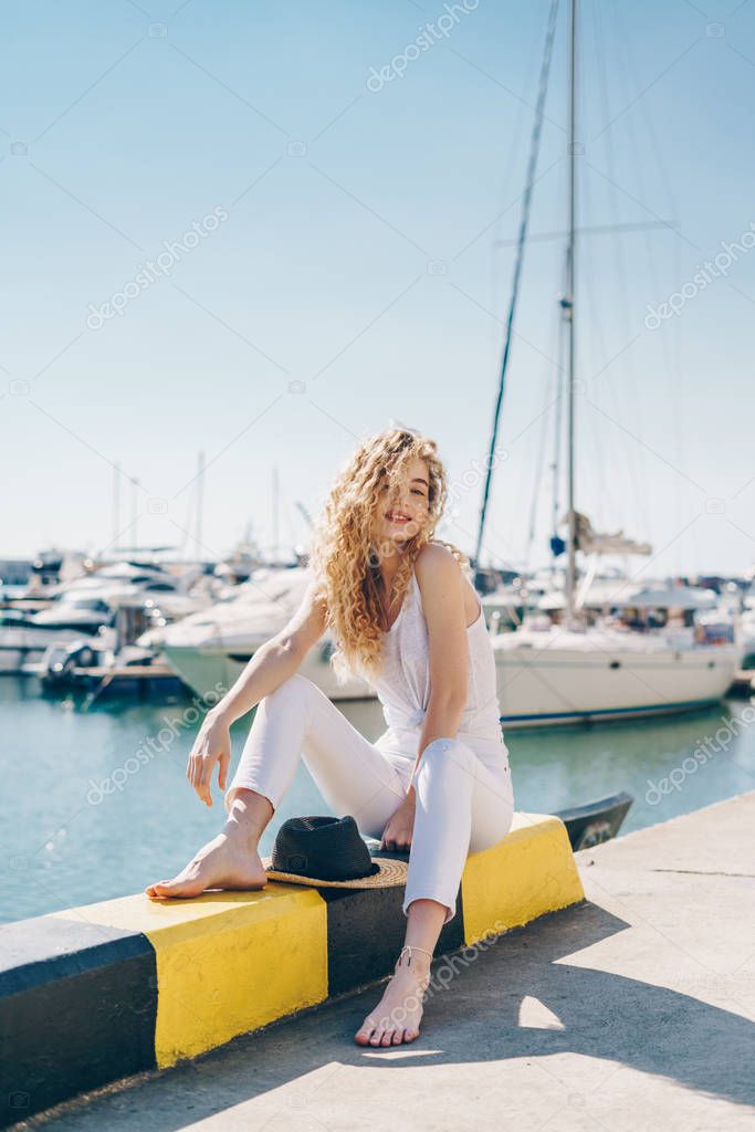 curly blonde with a smile in a relaxed pose sits on the parapet of the pier against the background of boats. bare feet
