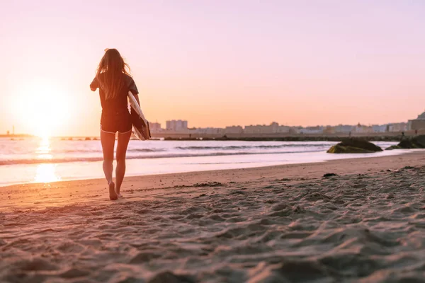 Sullo sfondo di un bellissimo tramonto, la ragazza va in lontananza lungo l'oceano nella sua tavola da surf mano — Foto Stock