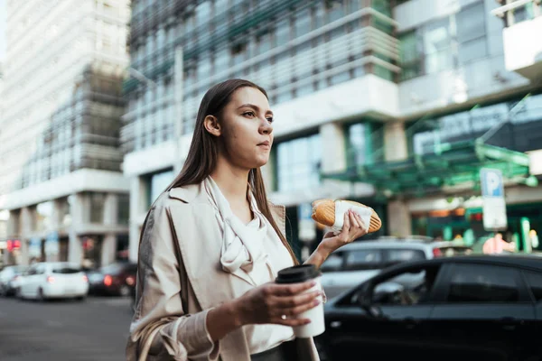 Девушка на улице задумчиво жует хот-дог и держит кофе — стоковое фото