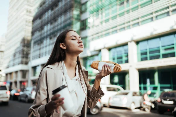 Девушка кусает сэндвич и с удовольствием закрывает глаза. фон здания — стоковое фото