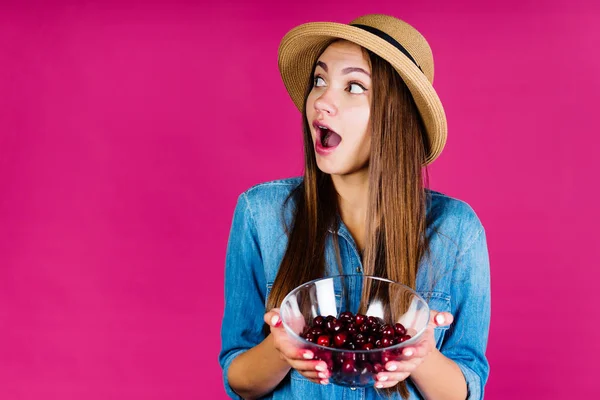 Fondo rosa. chica sorprendida, indignada de coquetería, abriendo la boca y entrecerrando los ojos, sostiene una taza con cerezas en la mano — Foto de Stock