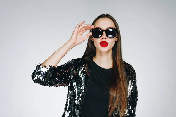 Una chica elegante con lápiz labial rojo en sus labios llenos y con una expresión de conciencia de su irresistibilidad se aferra a sus gafas negras. fondo gris claro — Foto de Stock