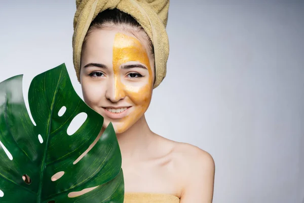 Aziatisch meisje met een geel masker op de helft van haar gezicht, een handdoek op haar hoofd, haar schouders zijn naakt, een monstera blad in haar hand. — Stockfoto