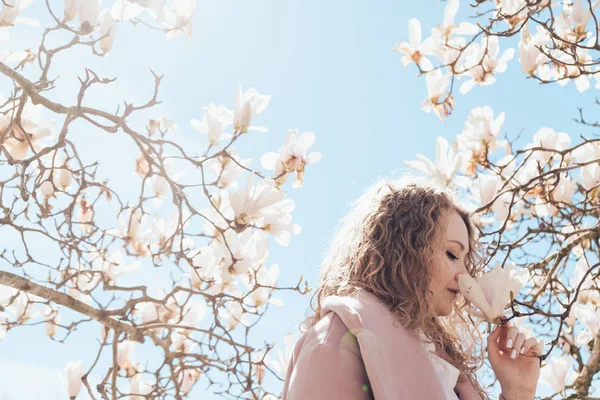 Rubia de placer inhala un aroma de magnolia flores de pie bajo un árbol — Foto de Stock