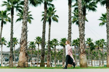 Palto giyen kız hızla Sochi parkındaki palmiye ağaçlarının arka planında yürüyor.