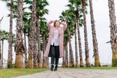 Patikadaki bir palmiye yolunda açık renk ceketli bir kız ellerini başının arkasında kaldırıyor.