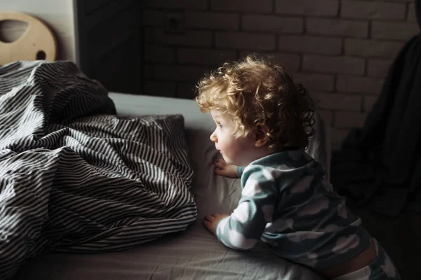 Bebé rubio rizado apoyado en la cama y mira hacia la ventana —  Fotos de Stock