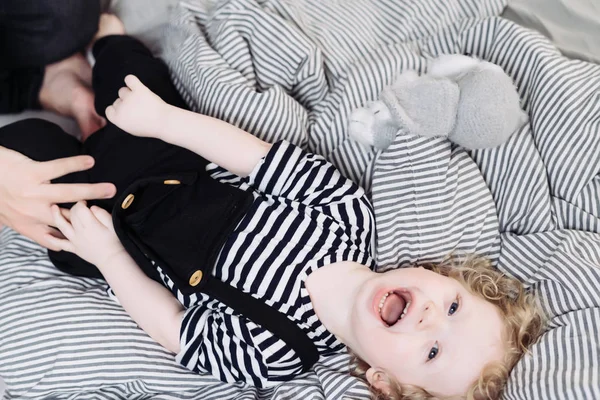 Curly angel baby laughs lying on the bed, hands put on his pants — Stock Photo, Image