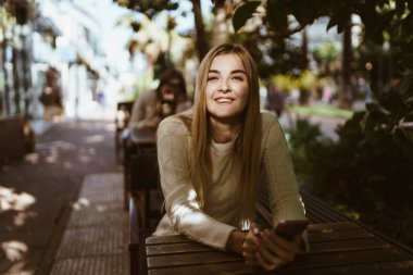 Uzun saçlı sarışın bir kafede oturur ve yüzünü aydınlatan güneşe gülümser.