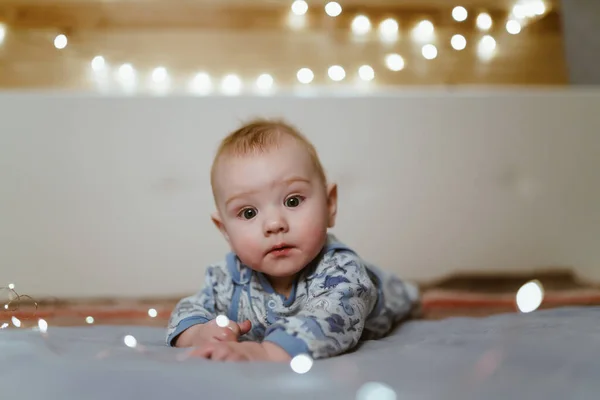 Un bebé maravilloso yace boca abajo y mira con su mirada gentil e ingenua, poniendo sus manos hacia adelante. alrededor de las bombillas — Foto de Stock