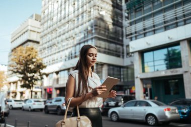 Kız iş merkezinin caddesinde yürüyor yol boyunca ve arabalar boyunca ve tablete bakıyor.