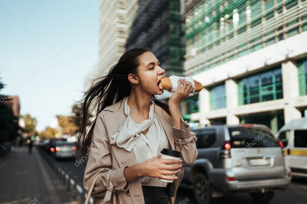 the wind blows the hair of a girl walking along the street of a business center and eagerly biting a bun with coffee in her hand