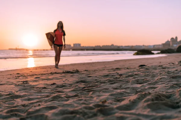 Sullo sfondo di un magnifico tramonto sull'oceano, una ragazza cammina con una tavola da surf sotto il braccio. Una grande città può essere vista in lontananza . — Foto Stock