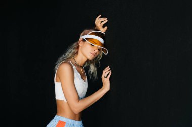 Siyah bir arka planda, güneşlik ve spor giysili bir kız başını duvara yasladı ve gözlerini kapattı.