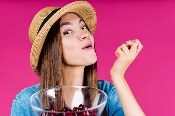 Pigg och förvånad leende flicka i en hatt äter körsbär från en vas i handen bakgrund rosa — Stockfoto