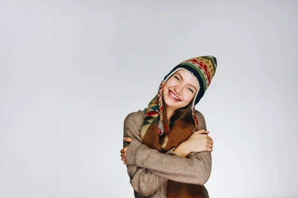 Joyfully broadly smiling girl in a warm colorful hat wrapped her arms around her shoulders background gray — Stock Photo, Image