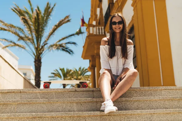 Un turista con i capelli lunghi in occhiali da sole sorridente con gioia si siede sui gradini della città meridionale della Spagna c'è uno skateboard nelle vicinanze — Foto Stock