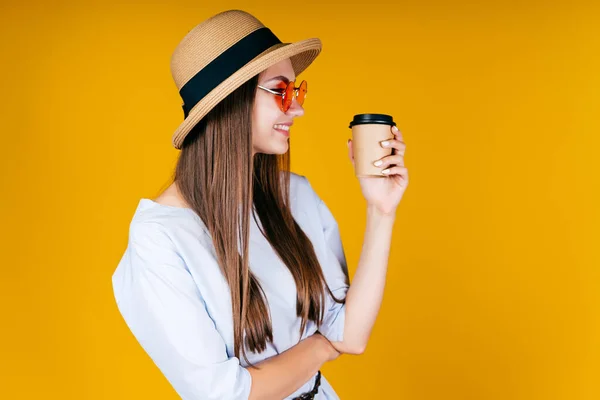 Ragazza elegante in un cappello e bicchieri tiene il caffè nelle sue mani e sorride guardando da qualche parte. sfondo giallo — Foto Stock