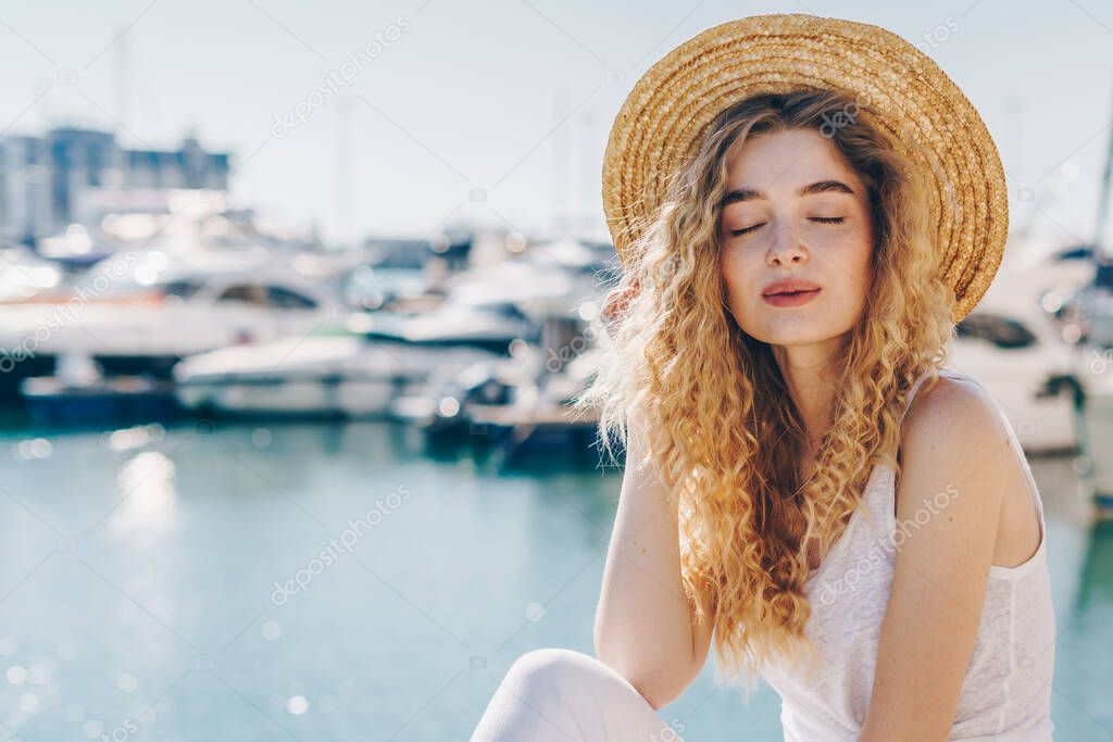 curly blonde with a restrained smile closing her eyes straightens her hat on the background of the pier