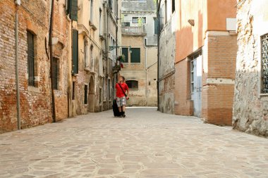 Yaz. İtalya. Venedik. Eski tuğla binalar arasında dar bir sokakta bavulla kırmızı bir gömlek ve siyah-beyaz kareli şort bir adam girer