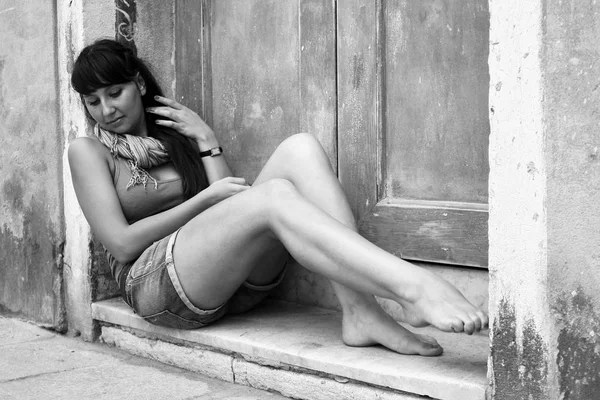 イタリア ヴェネツィア 素足でショート パンツとベストで長い黒髪の少女は古い木製のドアの横にある目の前に座っています 黒と白の写真 — ストック写真