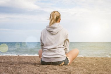 Sıcak, güneşli bir yaz günü. Gri kazaklı ve kot şortlu bir kız üzgün sahilde sırtı kameraya dönük oturur ve denize bakar.