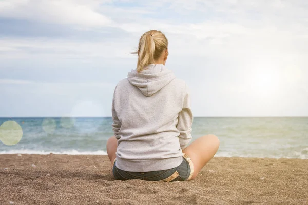 Sıcak, güneşli bir yaz günü. Gri kazaklı ve kot şortlu bir kız üzgün sahilde sırtı kameraya dönük oturur ve denize bakar.
