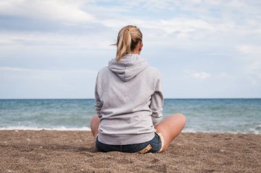 Sıcak, güneşli bir yaz günü. Gri kazaklı ve kot şortlu bir kız üzgün sahilde sırtı kameraya dönük oturur ve denize bakar.